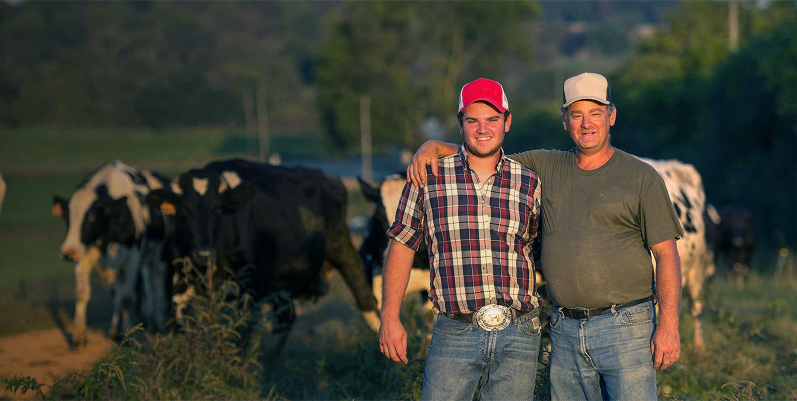 Two farmers standing in the pasture with cows in the background.