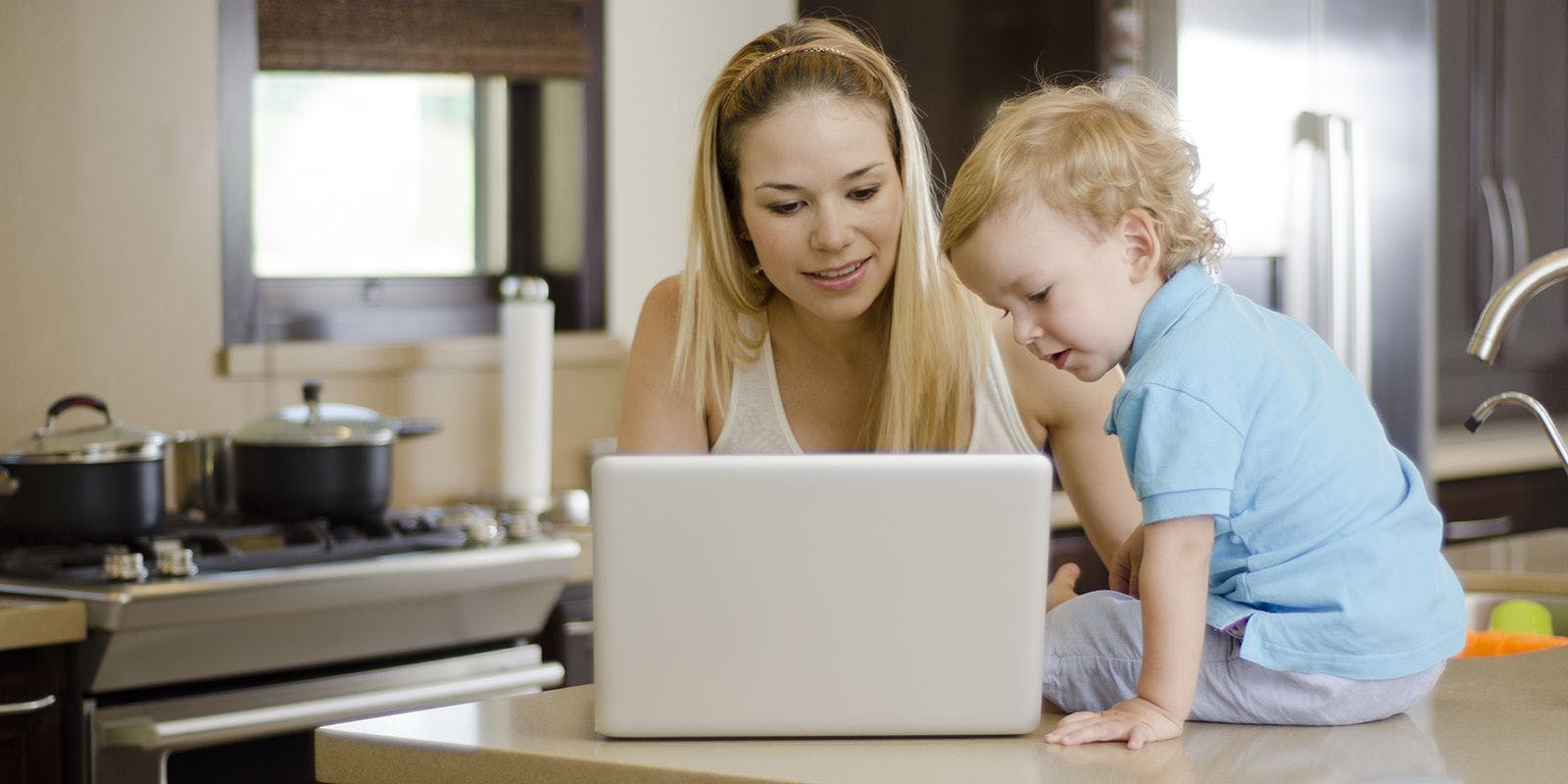 Blonde mom researches on her laptop while toddler sons sits on the counter in the kitchen.