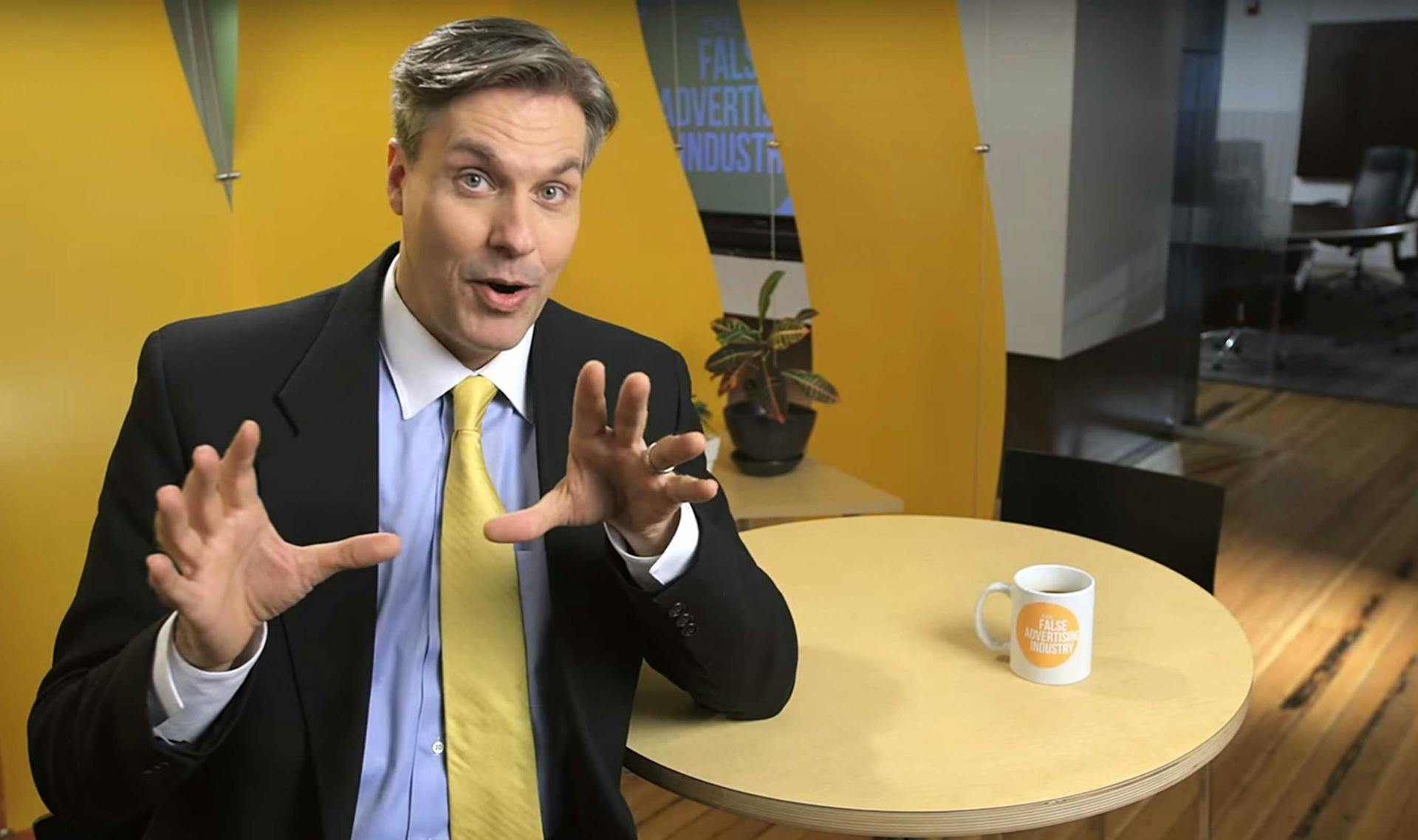 A man in a suit with his hands up with a mug on the table next to him reading "The False Advertising Industry."