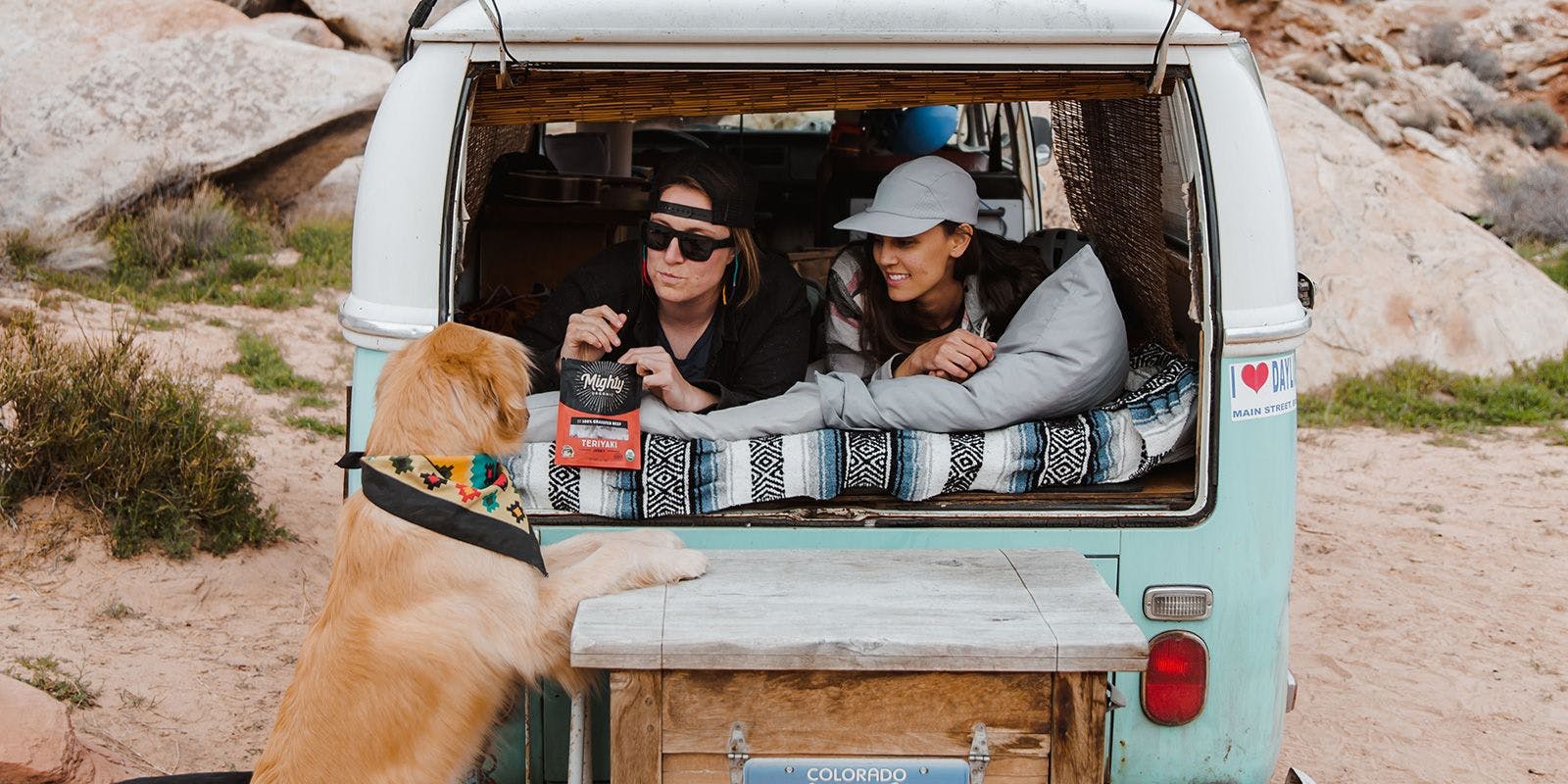 Two women lie in the back of a van on a beach eating Mighty Organic Jerky while their dog begs for a piece.