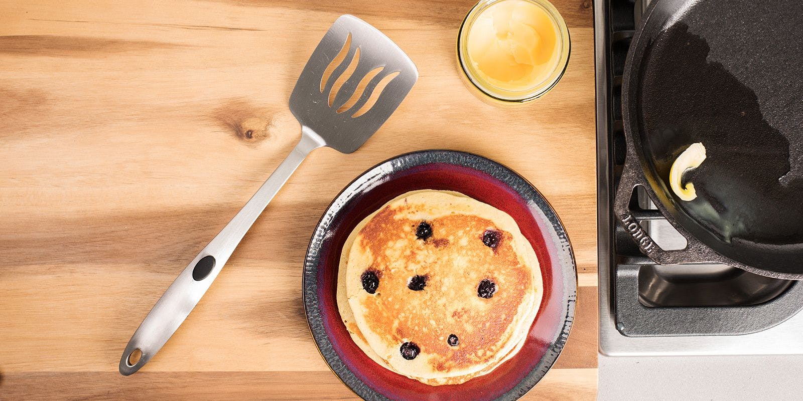 Fresh pancakes sit on a plate next to a jar of ghee and cooking spatula.