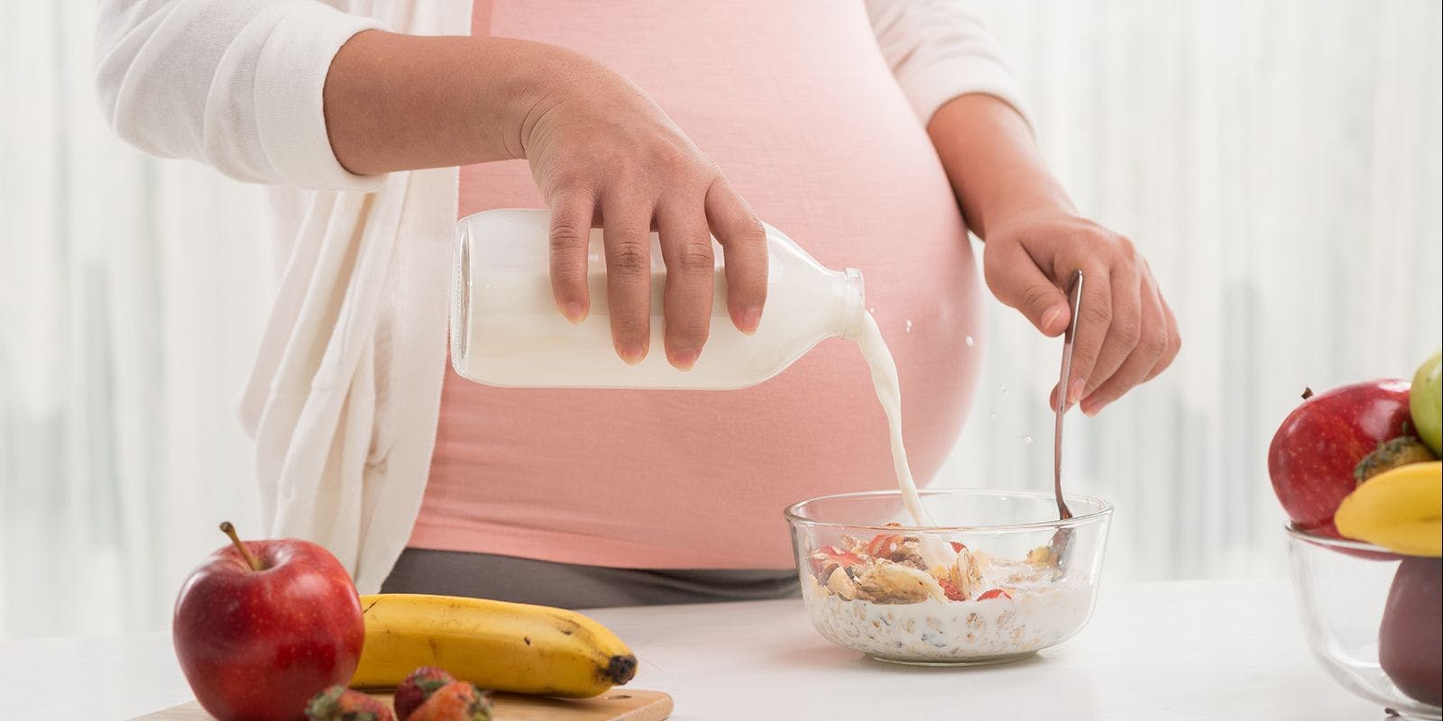 Pregnant woman pours milk into a bowl of cereal with fresh fruit.