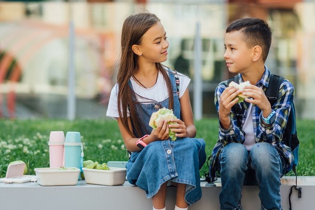 A girl and boy sit on a curb and eat lunch.