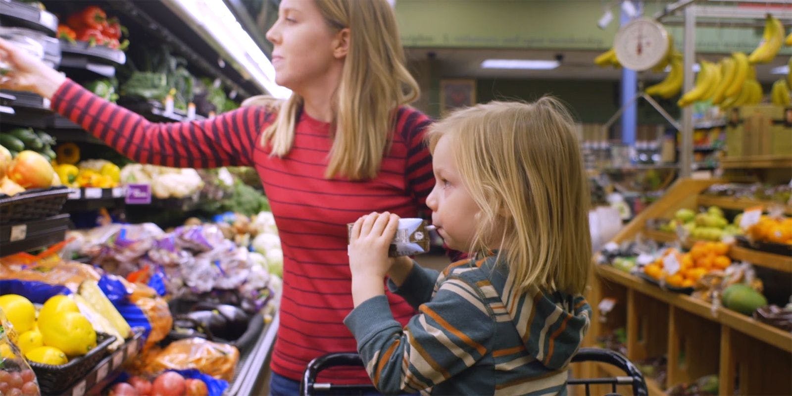 A woman shops in the produce aisle with a young child in the cart seat drinking chocolate milk.