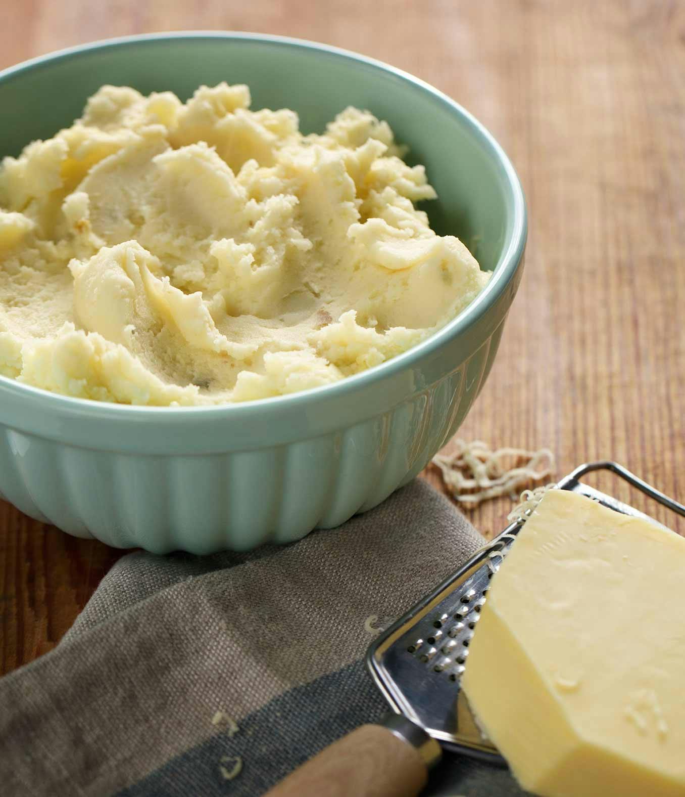 Raw cheddar and garlic mash potatoes in a large bowl with a block of cheddar cheese on the side.