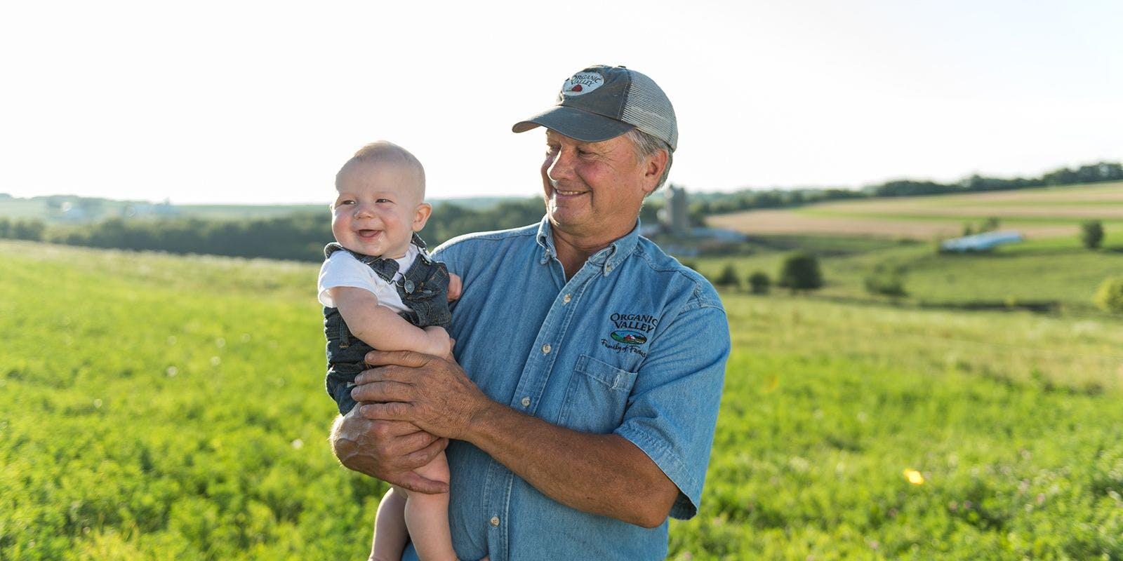 A farmer standing in a pasture holding his grandson who is smiling at something off camera.