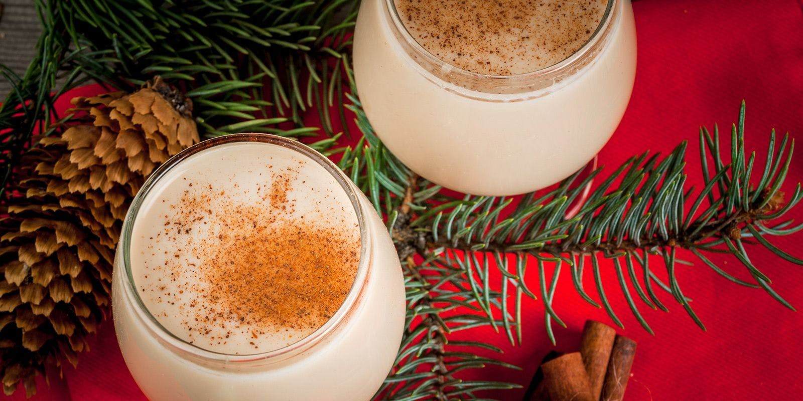 Glasses of eggnog dusted with nutmeg with evergreen branches, pine cones and cinnamon sticks around them on a red tablecloth.