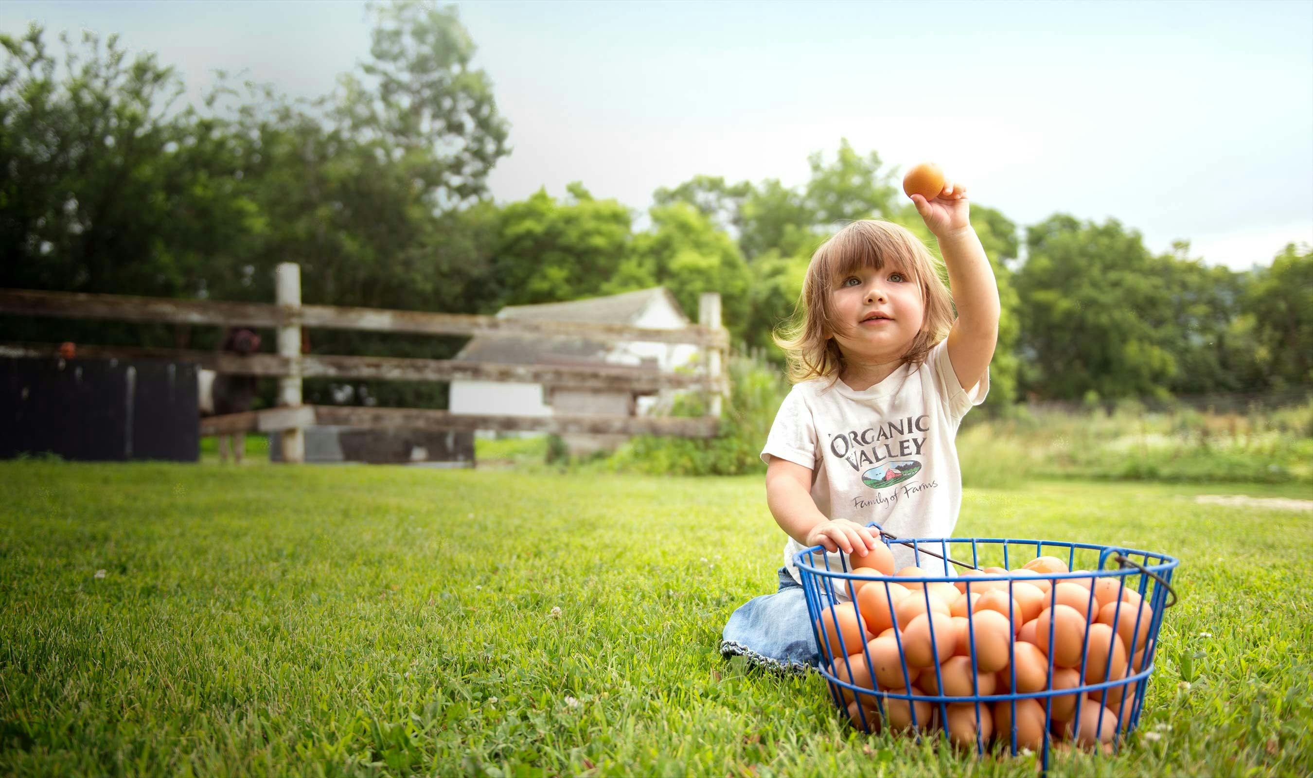 A girl holding an egg with a basket of eggs sitting in front of her.